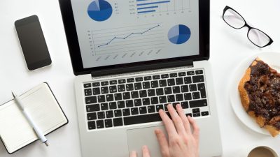 Woman hands working on a laptop with a diagram on it, glasses, mobile, office supplies, mug and a piece of pie on the plate. Business concept, lay flat